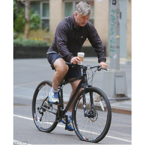 Alec Baldwin, riding a bike and carrying a takeaway coffee, looking suitably annoyed that his front tyre has gone flat
