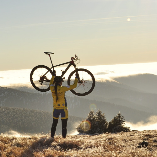 A woman standing on a mountain, holding a bicycle above her head, facing away from the camera