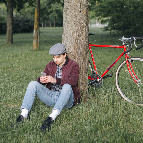A young man sitting against a tree, looking at his phone. A red bicycle leans on the tree behind him.