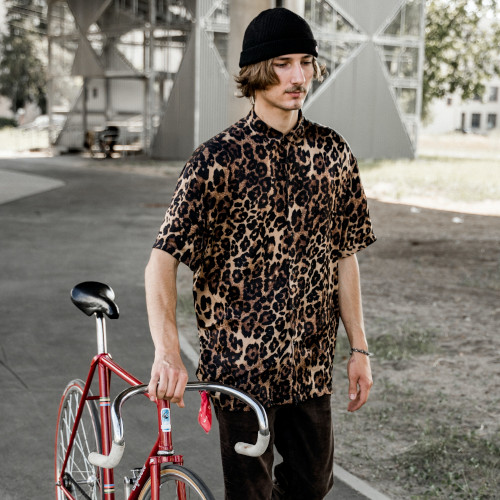 A young man in a leopard-print shirt and black beanie, wheeling a vintage bicycle