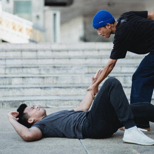 A guy on his back lying at the bottom of a staircase, clearly in pain. His friend is reaching out an arm to help him up.