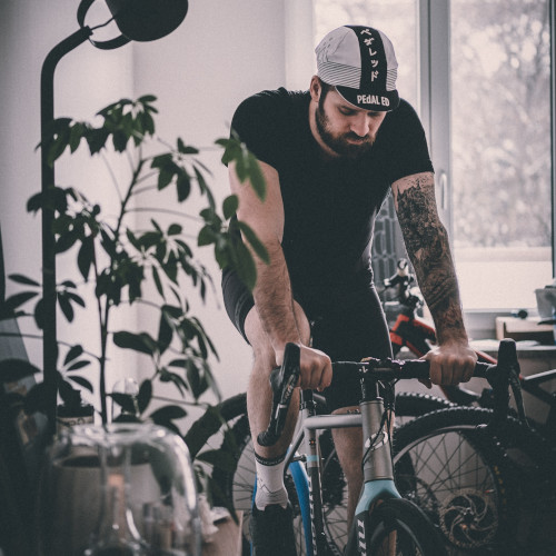 A bearded cyclist with a sleeve tattoo on an indoor trainer, next to a pot plant. He looks tired.