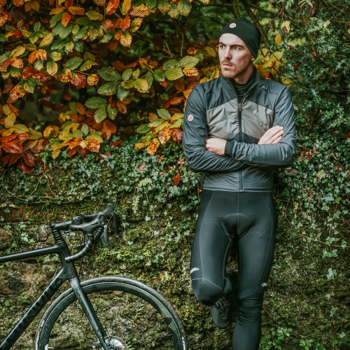 A lycra-clad cyclist leaning against a hedge with crossed arms, looking into the distance. A road bike is next to him.