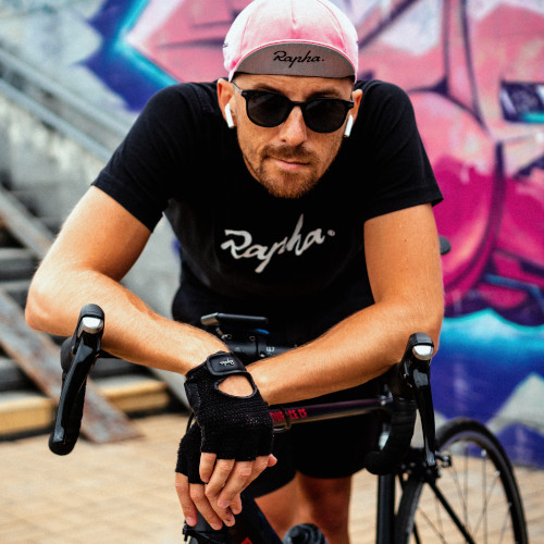 A cyclist with a black t-shirt and a pink cycling cap, leaning on his bike's handlebars in front of a graffitied wall