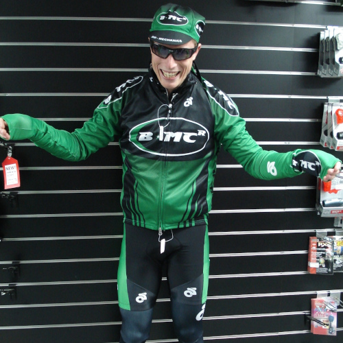 A cyclist dressed in multiple items of BMCR-branded clothing, standing against slatwall and looking like a fool