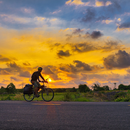 A cyclist riding along an empty road, a green field and sunset in the distance
