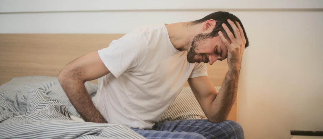 A man sitting on the side of an unmade bed, holding his head in apparent pain.
