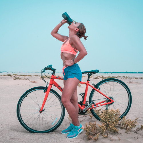 A woman standing next to a bicycle on a beach, drinking from an upturned water bottle