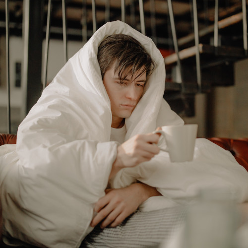 A man wrapped in a white quilt and holding a mug. He looks extremely unwell, and pale to the point of a consumptive Victorian-era child.