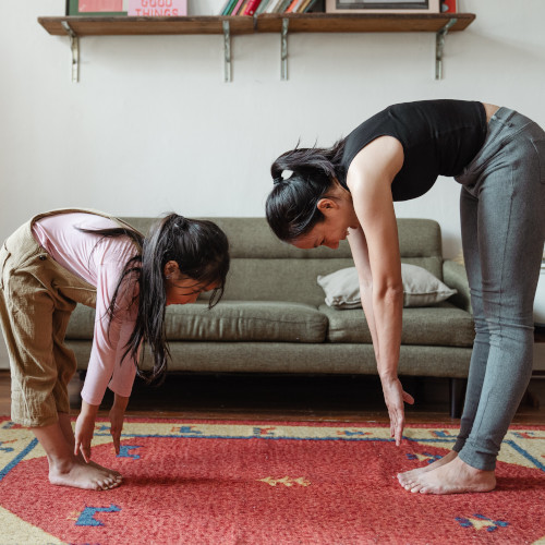 A mother and daughter face each other on a rug, both trying to touch their toes. Both are failing.