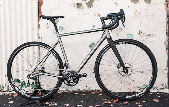 A custom-built Bossi Grit SX titanium bicycle against a white wall with peeling paint