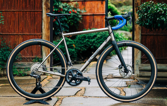 A custom-built Bossi Summit titanium road bike against a backdrop of wood and greenery. The blue handlebar tape really pops!