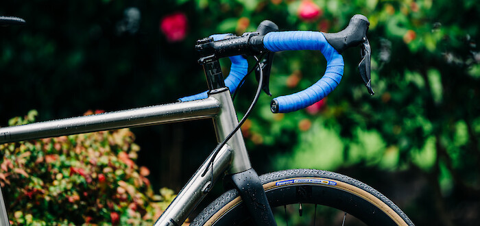 Partial side view of a custom-built Bossi Summit titanium road bike against a garden backdrop. The blue handlebar tape is vibrant against the greenery.