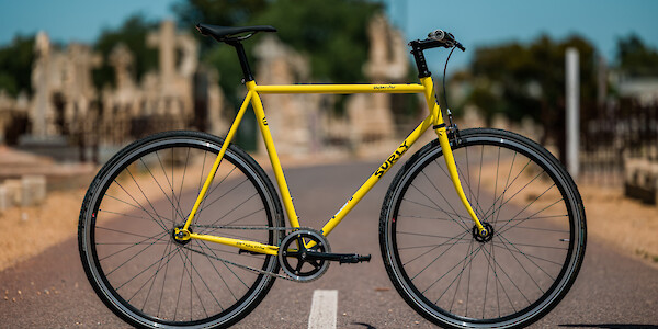 Surly Steamroller bicycle in Banana Candy yellow, custom-built, against a sunny graveyard backdrop