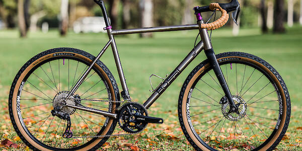 A custom-built Bossi Grit SX titanium bicycle with tan sidewall tyres, tan bar tape & purple accents, in a park setting