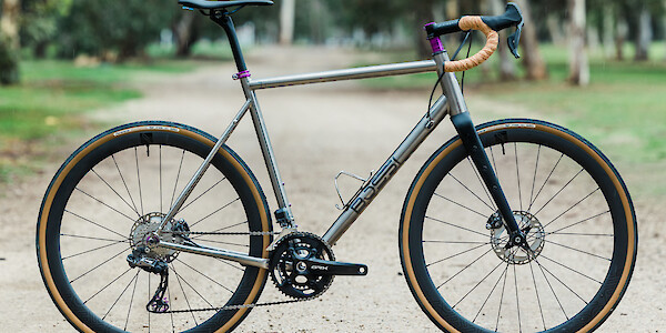A custom-built Bossi Grit SX titanium gravel bike with tan and purple components, on a flat gravel path