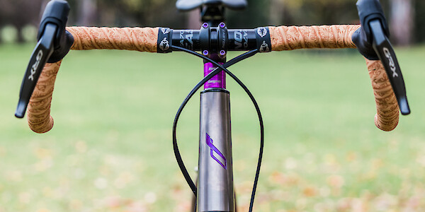 Headtube detail and handlebars on a custom Bossi Grit SX titanium gravel bike, showing the hand-painted head tube and contrasting purple/tan components