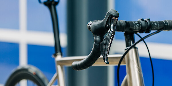 Right-hand shifter detail on a Bossi Strada titanium road bike in a custom build, viewed at a three-quarter angle, blue and silver panelling in the background