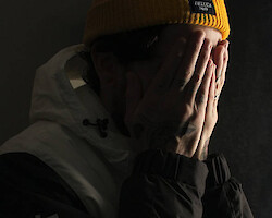 A man wearing a hoodie and yellow beanie, covering his face in despair