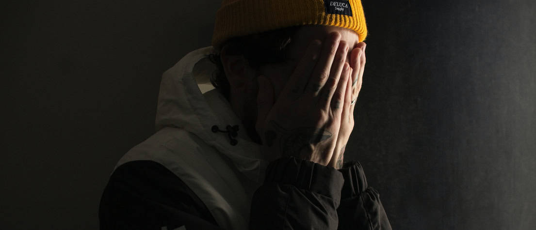 A man wearing a hoodie and yellow beanie, covering his face in despair