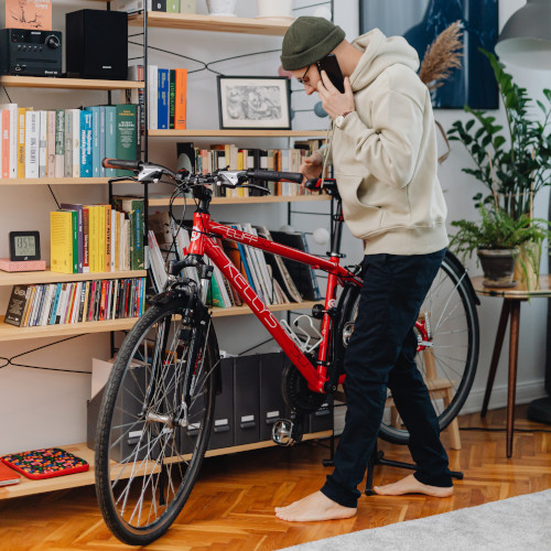 A young man in his living room, talking on the phone while looking at his bicycle.