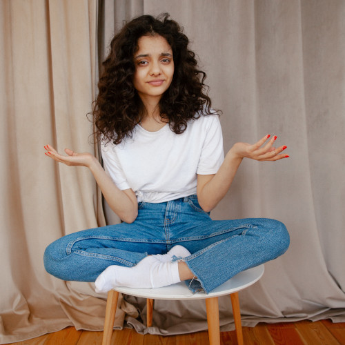 A young woman sitting cross-legged on a stool, holding her palms up quizzically