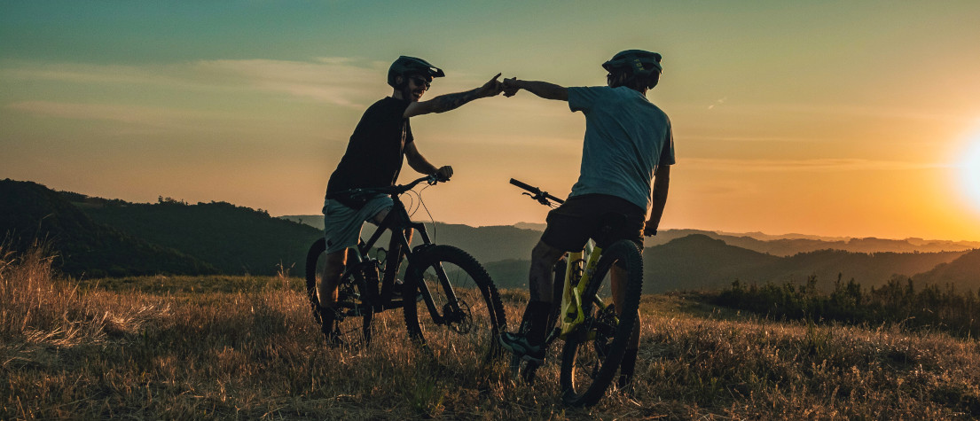 Two mountain bikers standing on a hill, bumping fists as the sun sets in the distance.