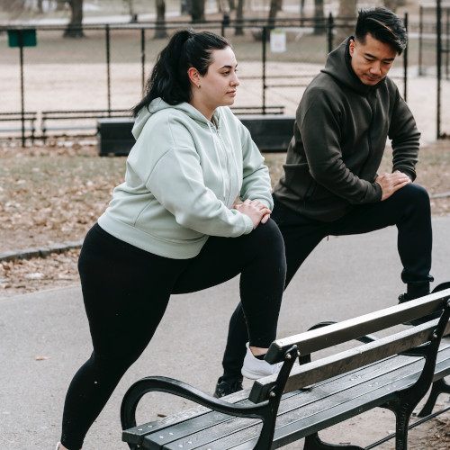A woman and a man in athletic gear, both stretching with their foot on a bench. They look distinctly unenthused.