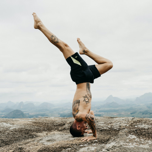 A tattooed, shirtless man doing a headstand on a rock. There are misty mountains in the distance.