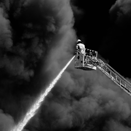 A firefighter on a raised platform, aiming a firehose down at plumes of smoke.