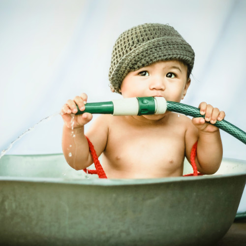 A baby wearing a knitted beanie and sitting in a shallow bowl. For some reason, he/she is attempting to eat a hose handle.