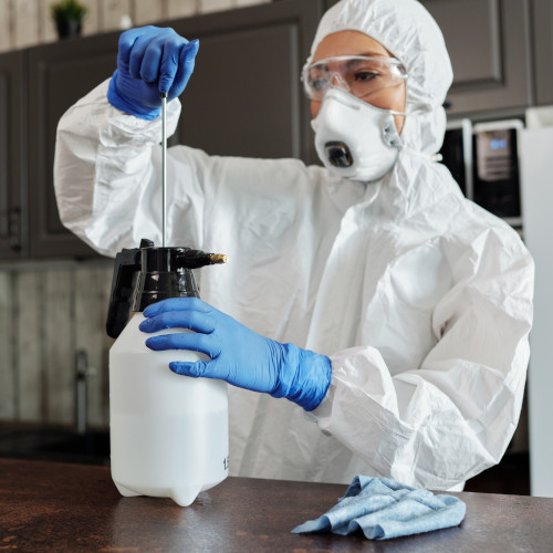 A woman in a kitchen, wearing a haz-mat suit and face mask, and preparing a spray bottle.