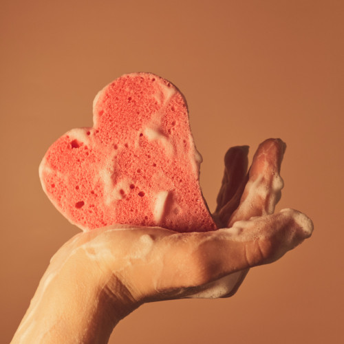 A hand holding a heart-shaped sponge, covered in soap suds.