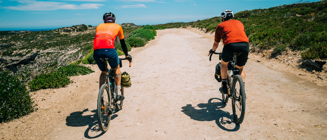 Two cyclists riding along a white gravel road, away from the camera. The sky is blue, their jerseys are bright orange.