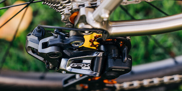 Detail of a Shimano GRX rear derailleur on a custom-built Bossi Grit SX titanium gravel bike, shot from above against a grassy backdrop