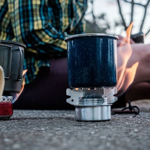 Close-up of a portable coffee-maker, a mug on top, flames licking out of the base. A bike rider can be seen sitting in the background, waiting for it to boil