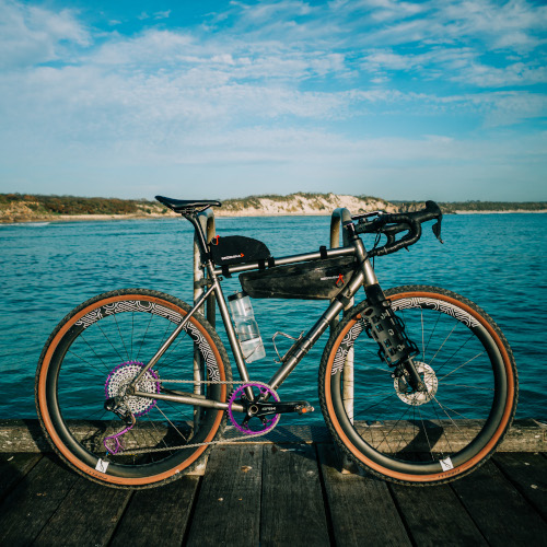 A Bossi titanium gravel bike - the Grit - leaning against a pole on a jetty, the sea behind it.