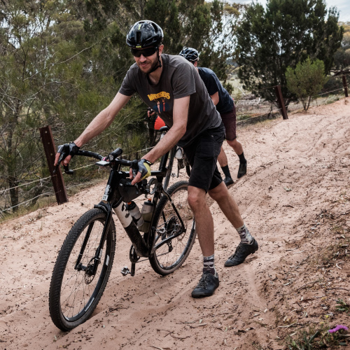 Two bikepacking cyclists, pushing their gravel bikes up a sandy slope.