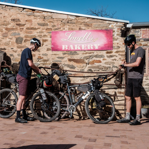 Two gravel riders packing their bikepacking bags outside a stone building on a sunny day. The building sign reads 'Lovell's Bakery'.