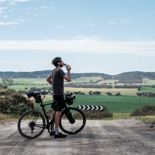 A gravel cyclist standing on a road, taking a drink of water, green fields in the horizon.