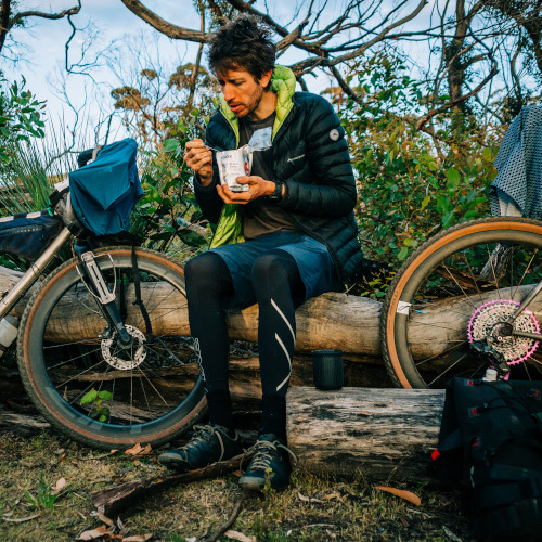 A gravel cyclist sitting on a log between two gravel bikes, eating a pre-prepared meal and taking a break on his bikepacking trip.