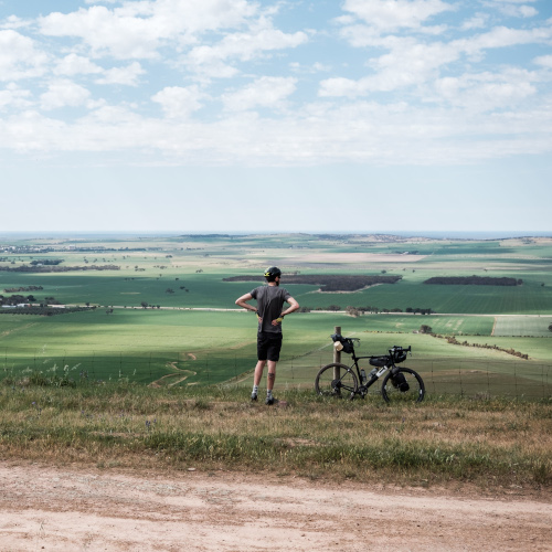 A gravel cyclist stopped on a ridge, looking out over a horizon of green fields. His gravel bike is next to him, leaning on the fence.