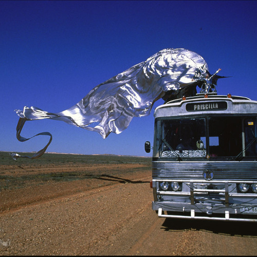 Still of Guy Pearce from the movie Priscilla, atop a bus, silver lame billowing behind him.