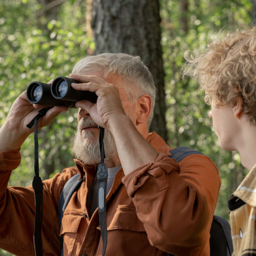 A bearded man in an orange shirt stands in the woods, looking through a pair of binoculars. A younger man is next to him, looking at him.