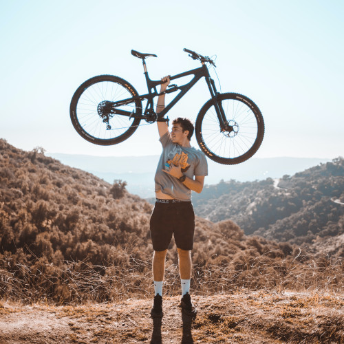 A young man stands on a hillside, lifting his mountain bike above his head in a show of triumph.