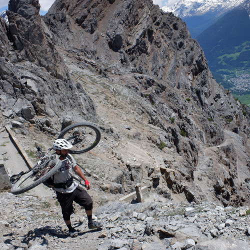 A man climbing out of a rocky valley, his mountain bike slung over his shoulder. The terrain looks hot and dry.