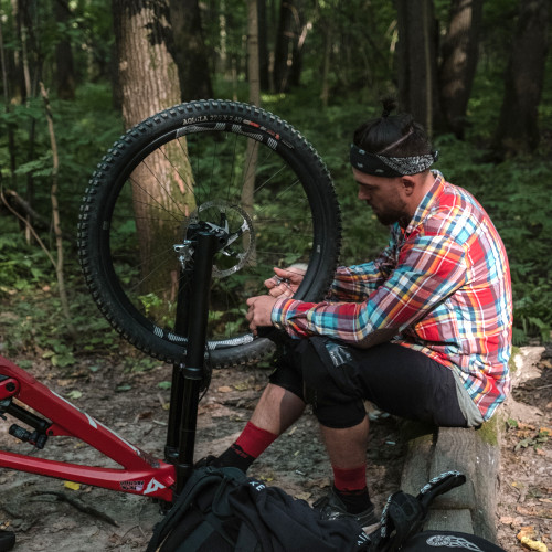 A mountain biker sitting on a log, fixing the spokes on his mountain bike wheel.