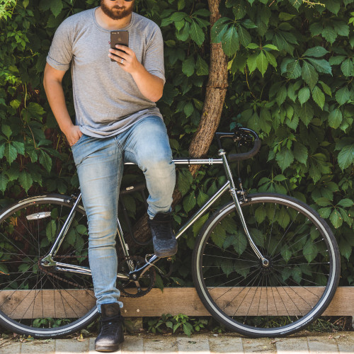 A man leans against a bike, looking at his phone, a leafy wall just behind him.