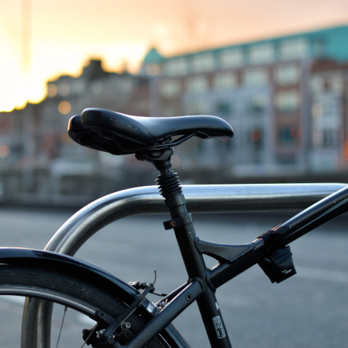 Partial frame detail of a basic black commuter bicycle next to a bike rack, city buildings in the background