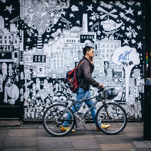 A man walking a bicycle past a black and white mural. He's on the phone, wearing a backpack.